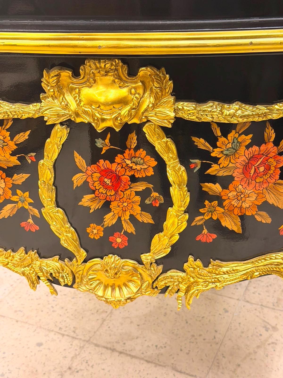 May include: A black lacquered furniture piece featuring ornate gold metal trim and relief carvings. The surface is decorated with hand-painted floral motifs in shades of orange, red, and yellow, framed by golden laurel wreaths and scrollwork.