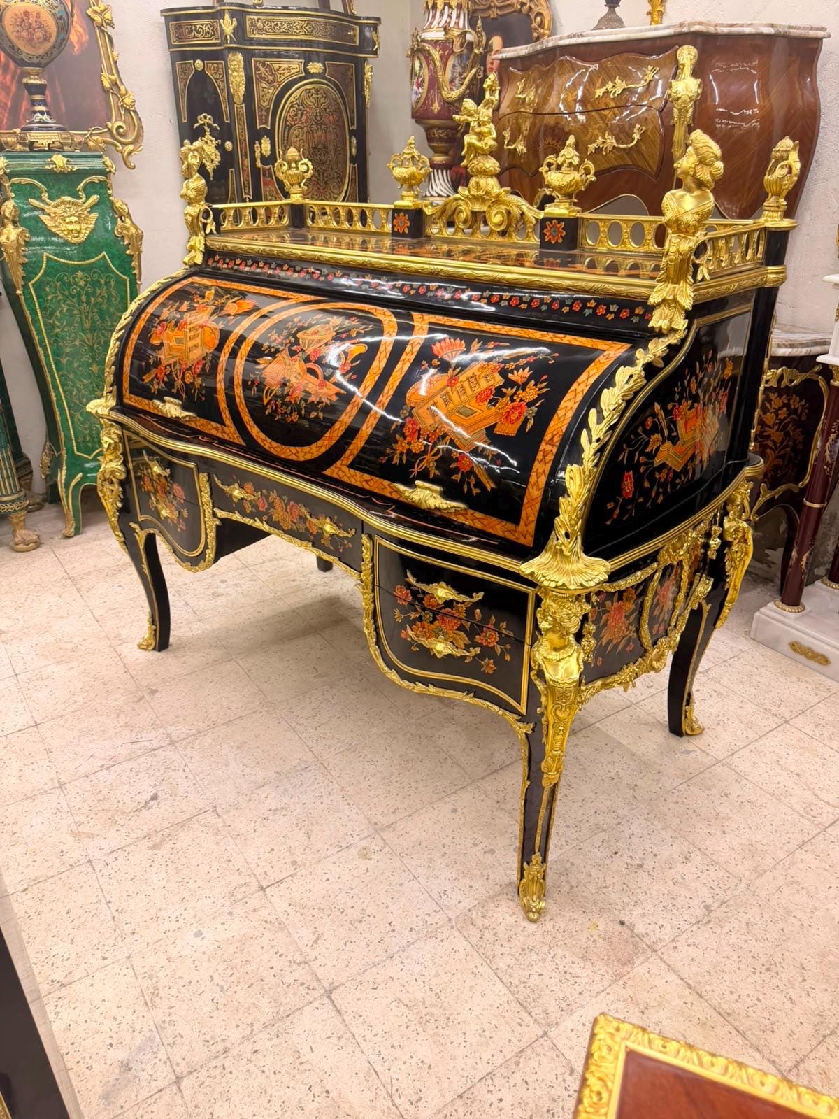 May include: A black lacquer roll-top desk featuring intricate floral marquetry in orange and gold tones. The piece is adorned with ornate gold-colored metal mounts, including figural accents on the corners and a decorative gallery rail.