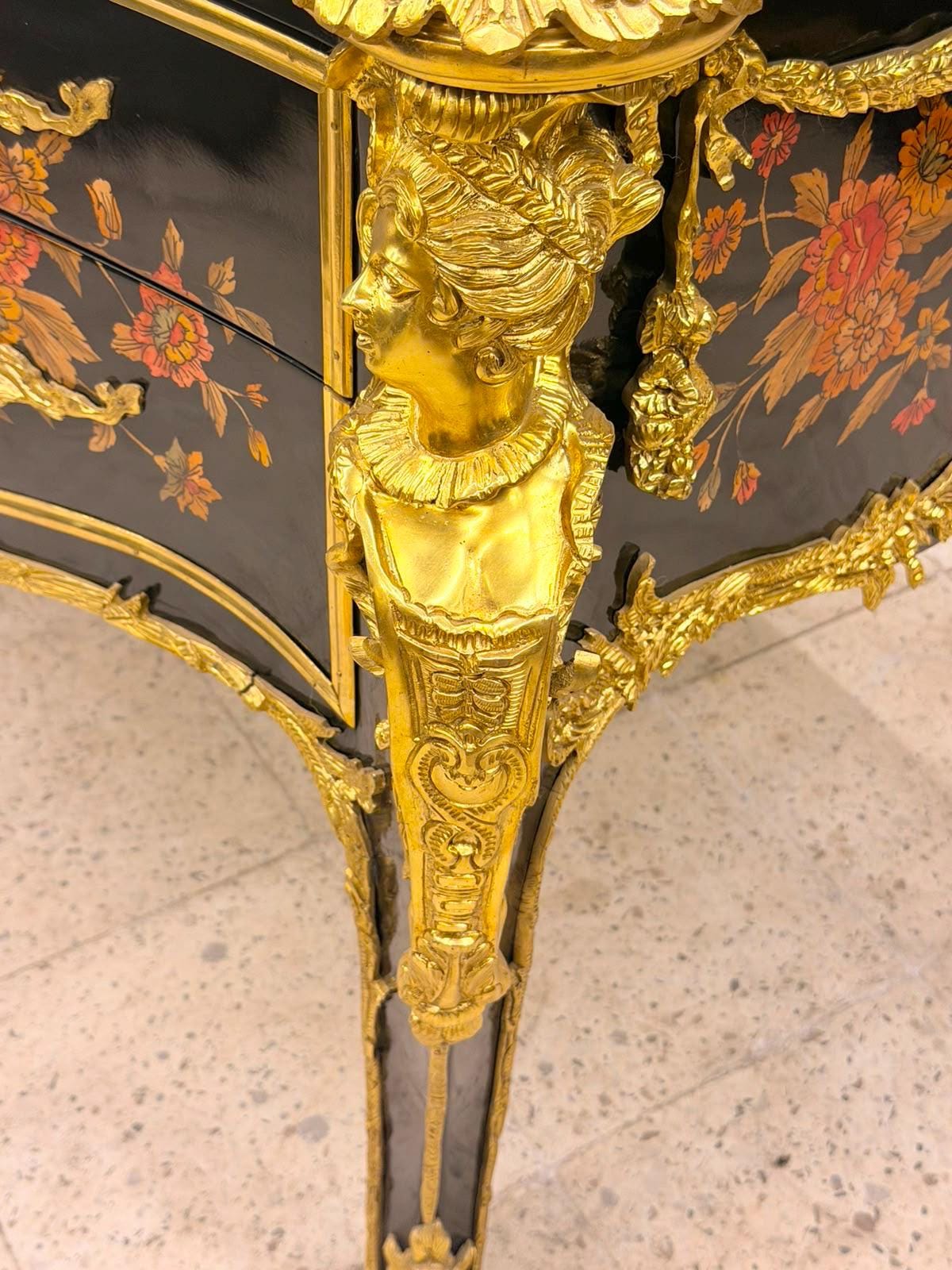 May include: A black lacquered chest of drawers featuring intricate floral patterns in shades of orange and green. The piece is adorned with ornate gold-colored metal mounts, including a prominent sculpted female bust on the corner leg.
