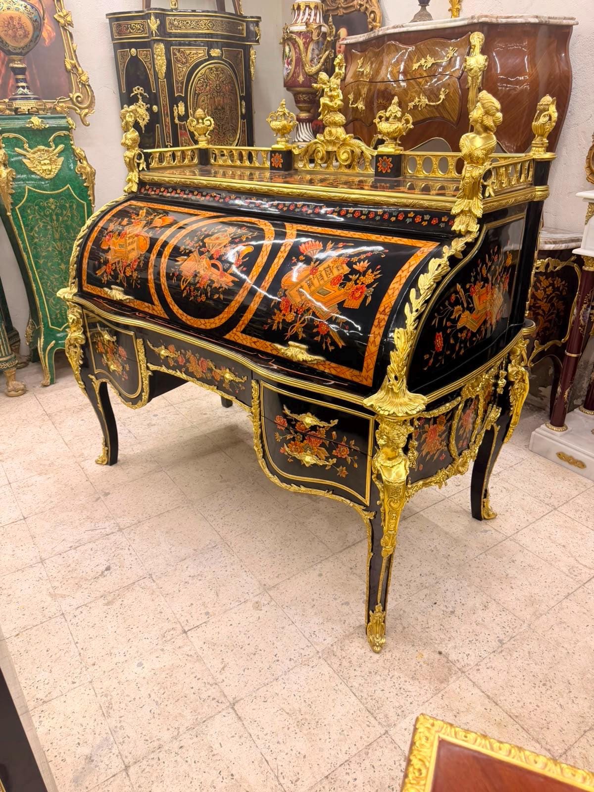 May include: A black roll-top desk featuring intricate gold-toned metal ornamentation, including ornate figures and scrolling foliage. The desk surface and drawers are decorated with colorful floral marquetry in shades of orange, red, and green.