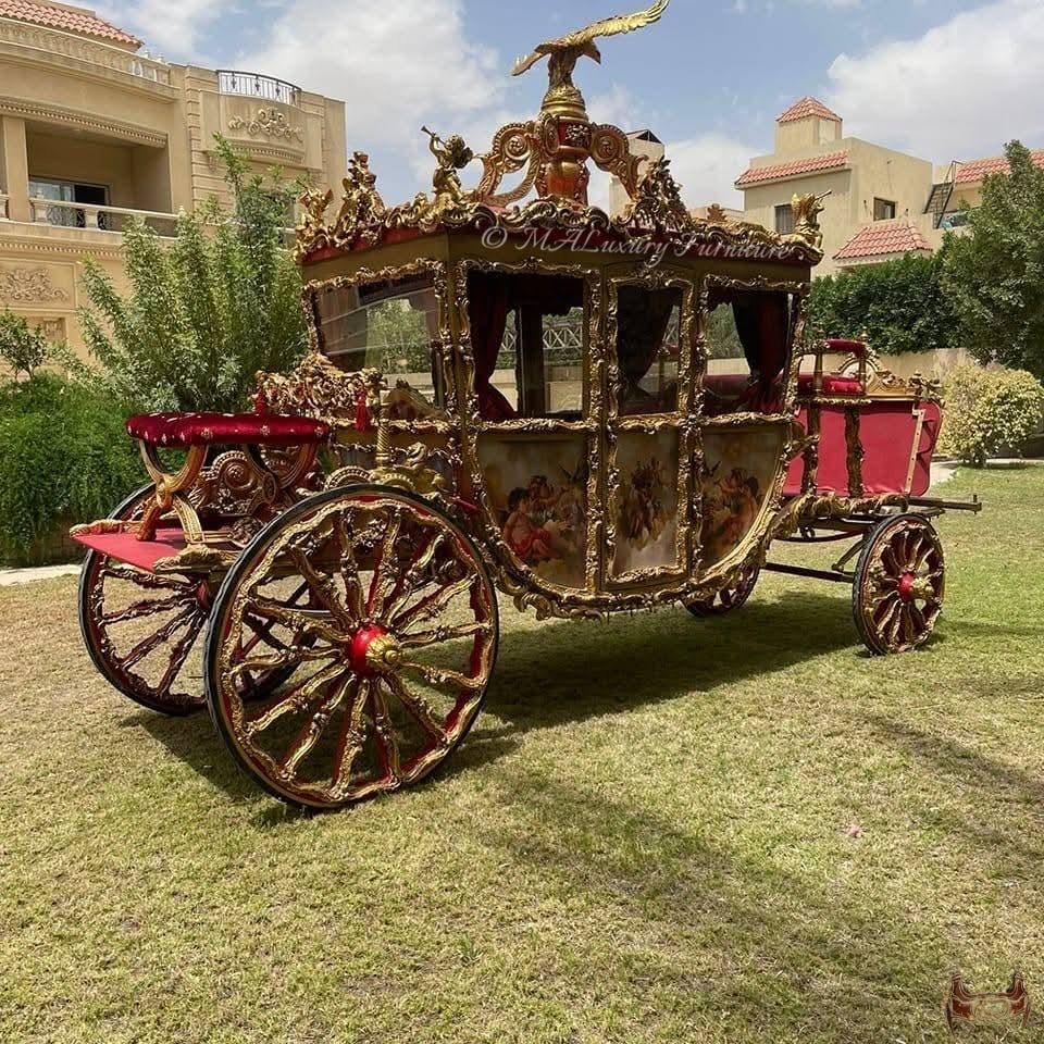 May include: A large, ornate horse-drawn carriage featuring intricate gold-painted carvings, red velvet upholstery, and painted panels depicting figures. The structure is topped with a golden eagle sculpture and includes large spoked wheels.
