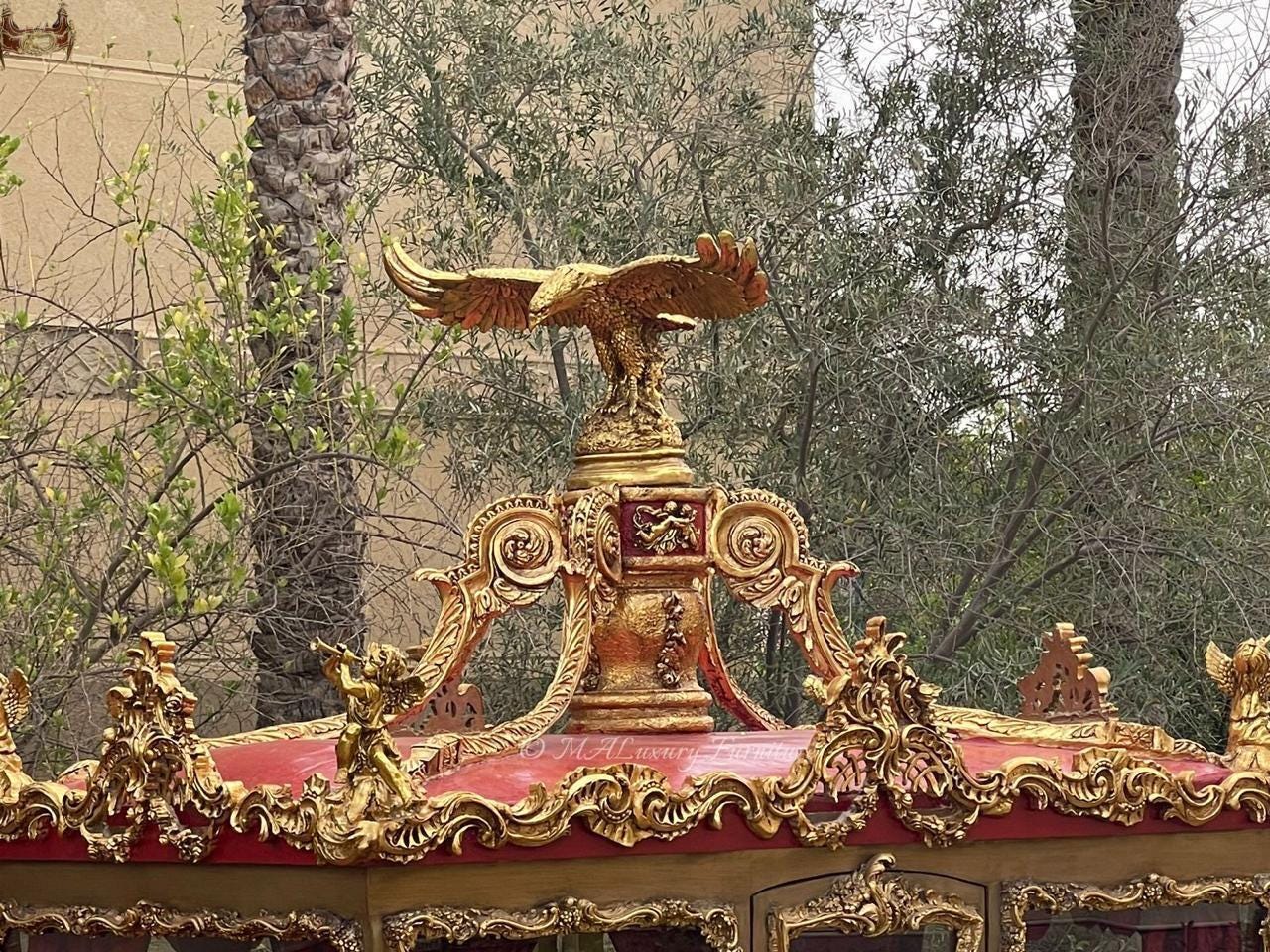 May include: A gold-colored eagle sculpture perched atop an ornate, red-roofed structure featuring intricate gold scrollwork, carved cherub figures, and a central pedestal. The structure displays detailed relief patterns and a deep red finish.