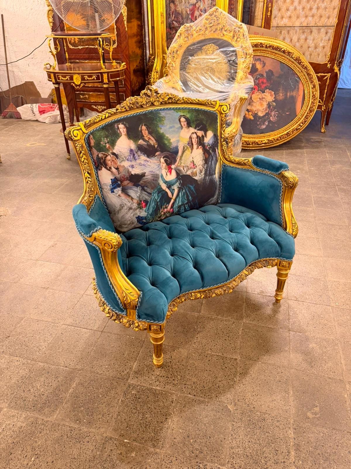May include: A teal velvet settee with a tufted seat and a gold-painted wooden frame. The backrest features a printed fabric panel depicting a group of women in historical attire. The piece has curved armrests and carved legs with gold detailing.