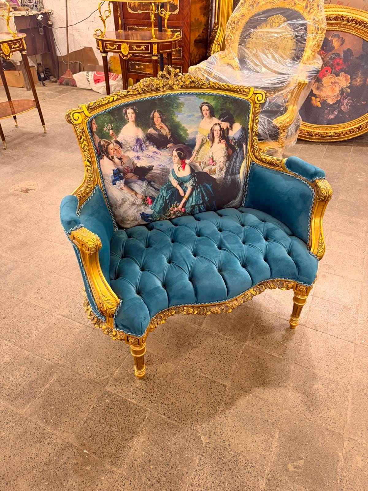 May include: A gold-framed armchair with a tufted teal velvet seat and a backrest featuring a printed scene of several women in historical attire. The wooden frame is carved with ornate details and finished in a metallic gold paint.
