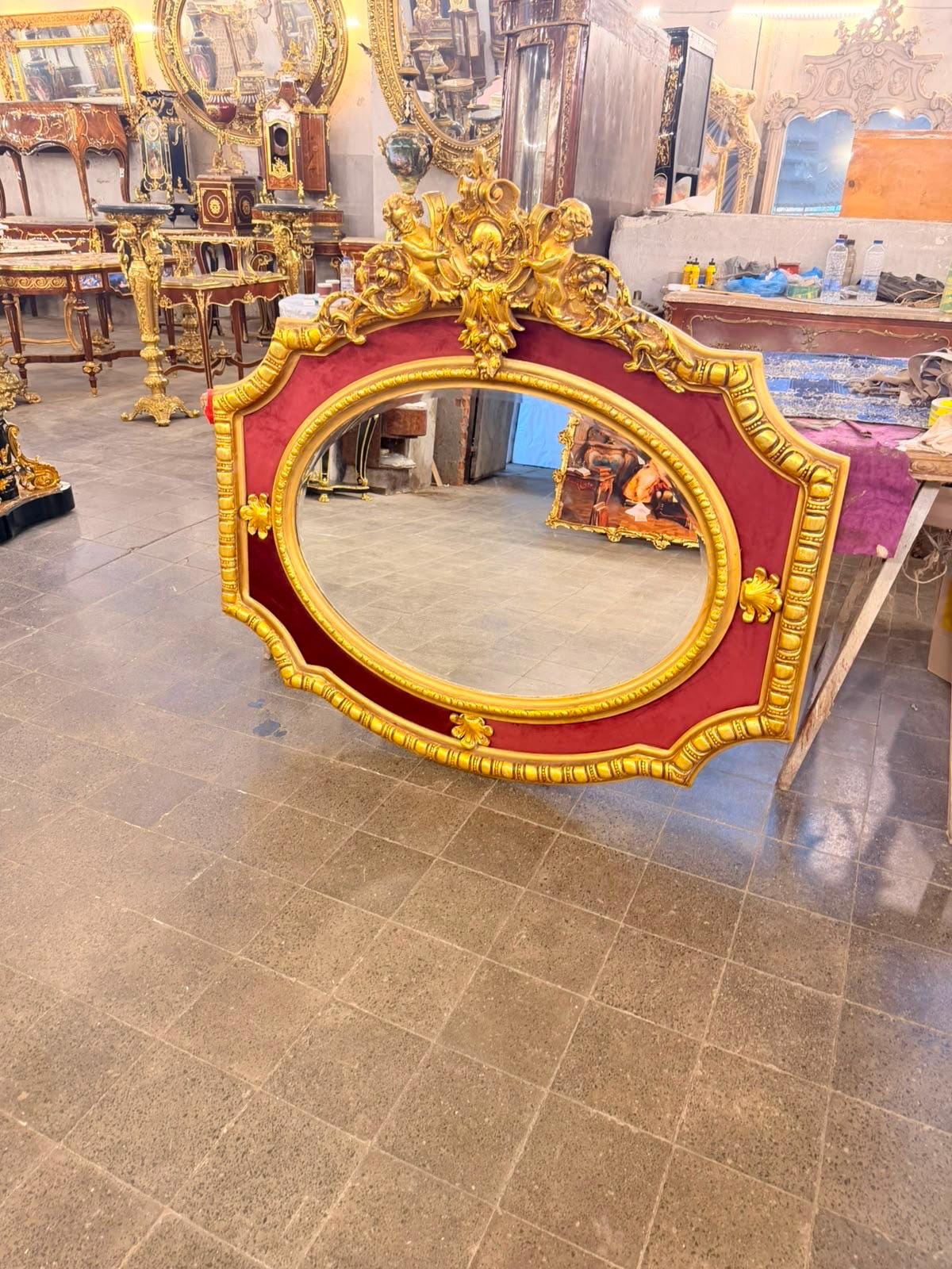 May include: An oval wall mirror featuring a wide, ornate gold-painted frame with intricate carved details and cherub figures at the top. The frame is accented with a deep red velvet-like border surrounding the central glass pane.