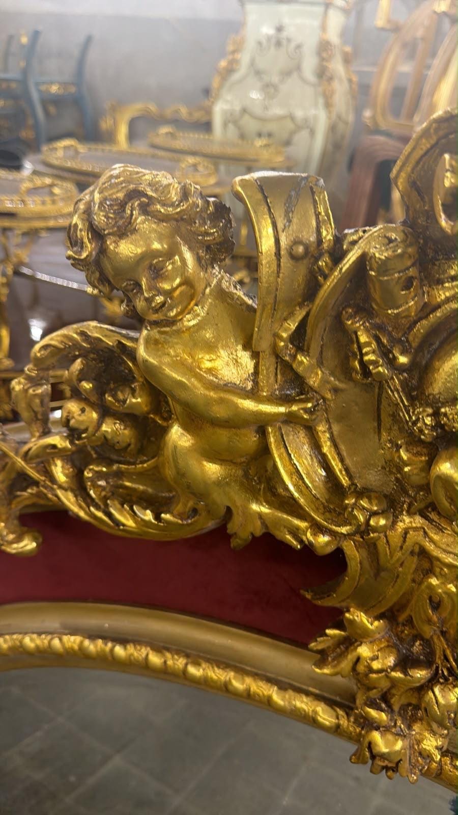 May include: A gold-colored decorative frame featuring a sculpted cherub figure in relief. The cherub is positioned next to a curved element, with ornate scrollwork and foliage details surrounding the figure. The frame has a deep red velvet border.