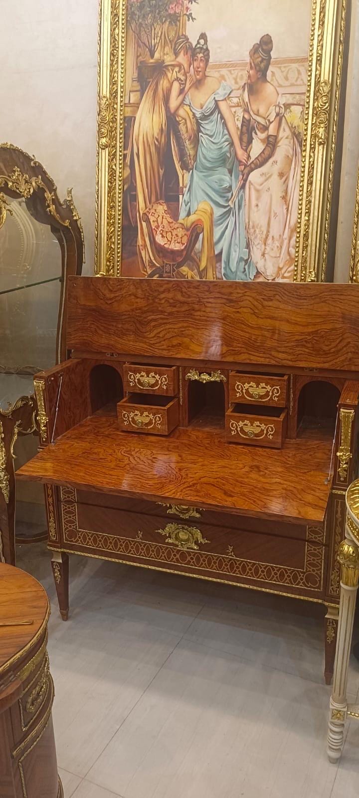May include: A wooden drop-front secretary desk with a polished burl wood finish, featuring gold-toned metal hardware, two small open drawers, and a large lower drawer. The desk has ornate gold metal accents on the legs and frame edges.