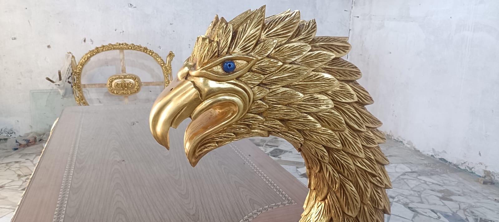 May include: A gold-colored eagle head sculpture featuring intricate feather detailing and a single bright blue eye. The sculpture is crafted with a smooth, curved beak and a textured neck, showcasing a metallic finish that reflects light across its surface.