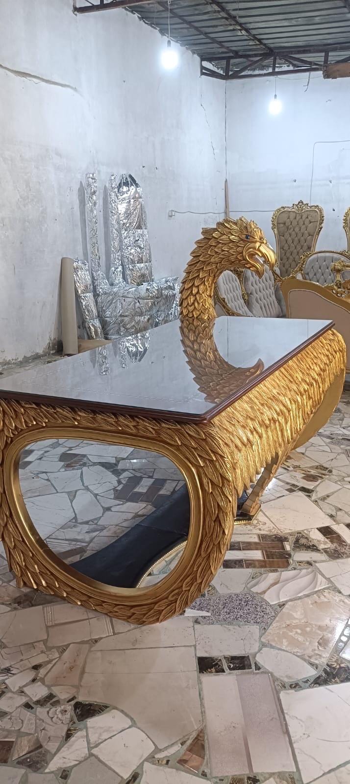 May include: A rectangular desk with a smooth glass top and a gold-colored base featuring intricate carved feather textures. The desk includes a prominent, sculpted eagle head extending from one side, with detailed plumage and a metallic gold finish.