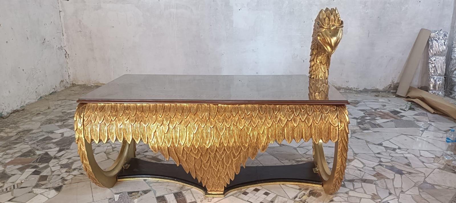 May include: A rectangular coffee table featuring a dark brown glass top and a gold-finished base intricately carved with a dense pattern of overlapping feathers. One corner of the table base extends upward into a sculpted golden eagle head figure.