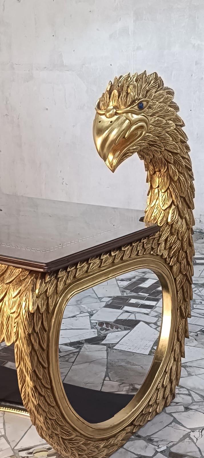 May include: A gold-colored table featuring a carved eagle head and neck as a support structure. The eagle has detailed feather textures and a small blue eye. The table has a rectangular dark brown top with a smooth surface and a rounded frame.