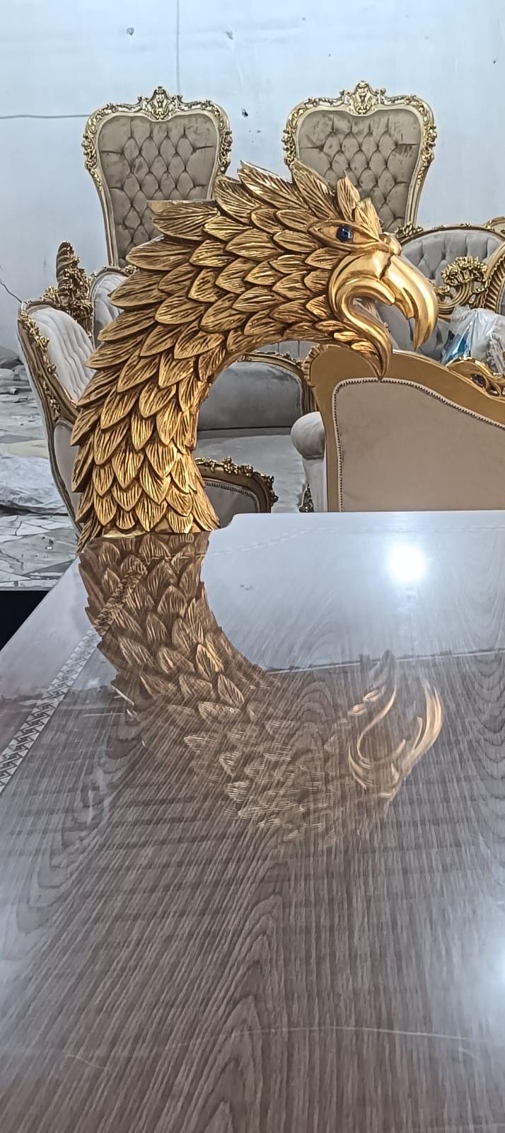 May include: A gold-colored eagle head sculpture with detailed feather textures and a dark eye, positioned on a reflective surface. In the background, several upholstered chairs with gold frames and tufted beige fabric are visible in the room.