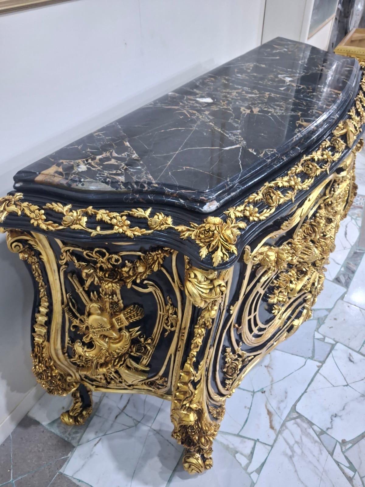 May include: A black chest of drawers featuring a polished black marble top with white veining. The exterior is decorated with intricate gold-colored metal relief carvings of floral motifs and ornate patterns, supported by four carved gold-colored feet.