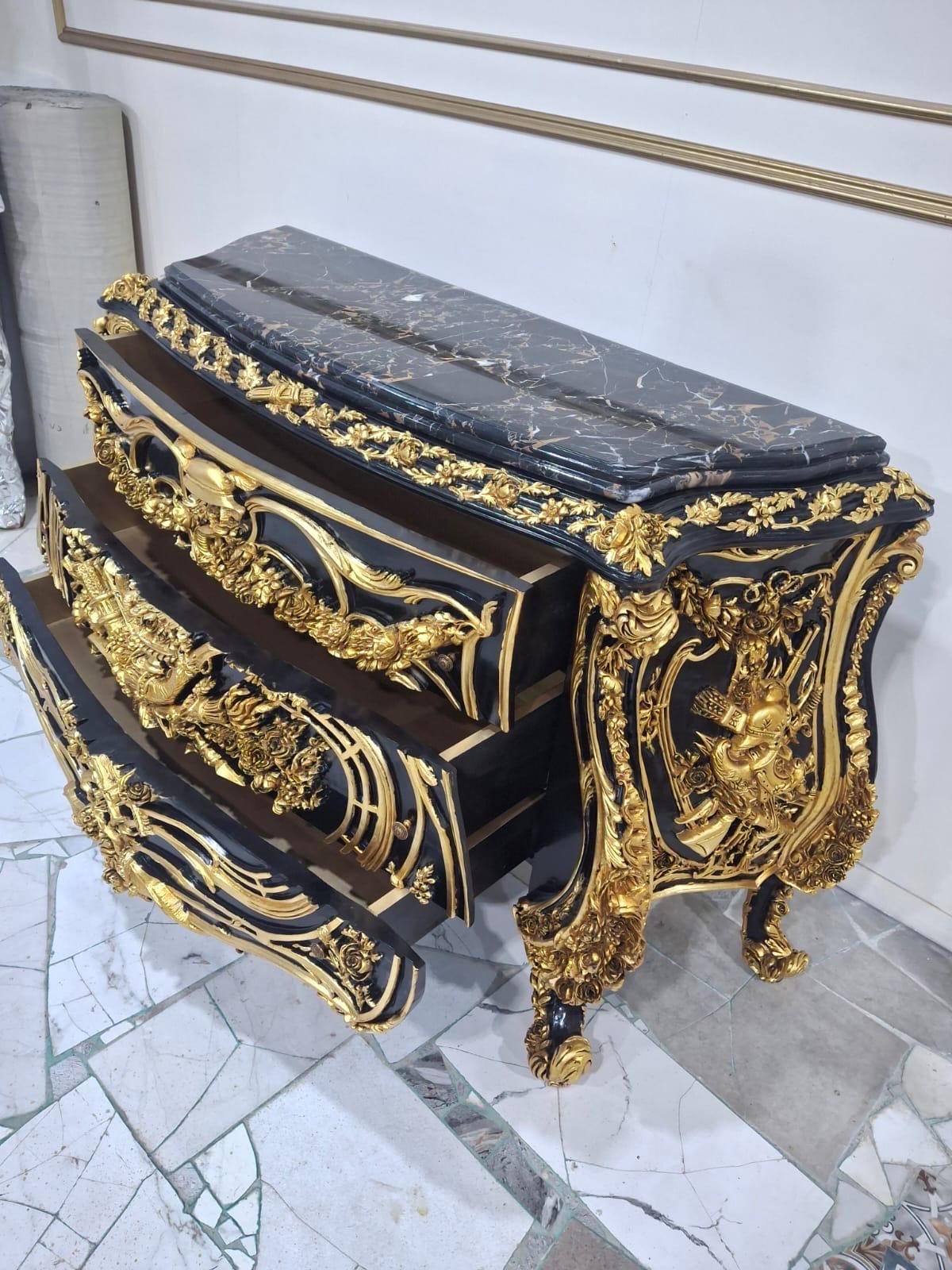 May include: A black wooden chest of drawers featuring a dark marble top and intricate gold-colored carved floral and scrollwork ornamentation. The piece has three pull-out drawers with curved fronts and ornate gold detailing on the side panels.