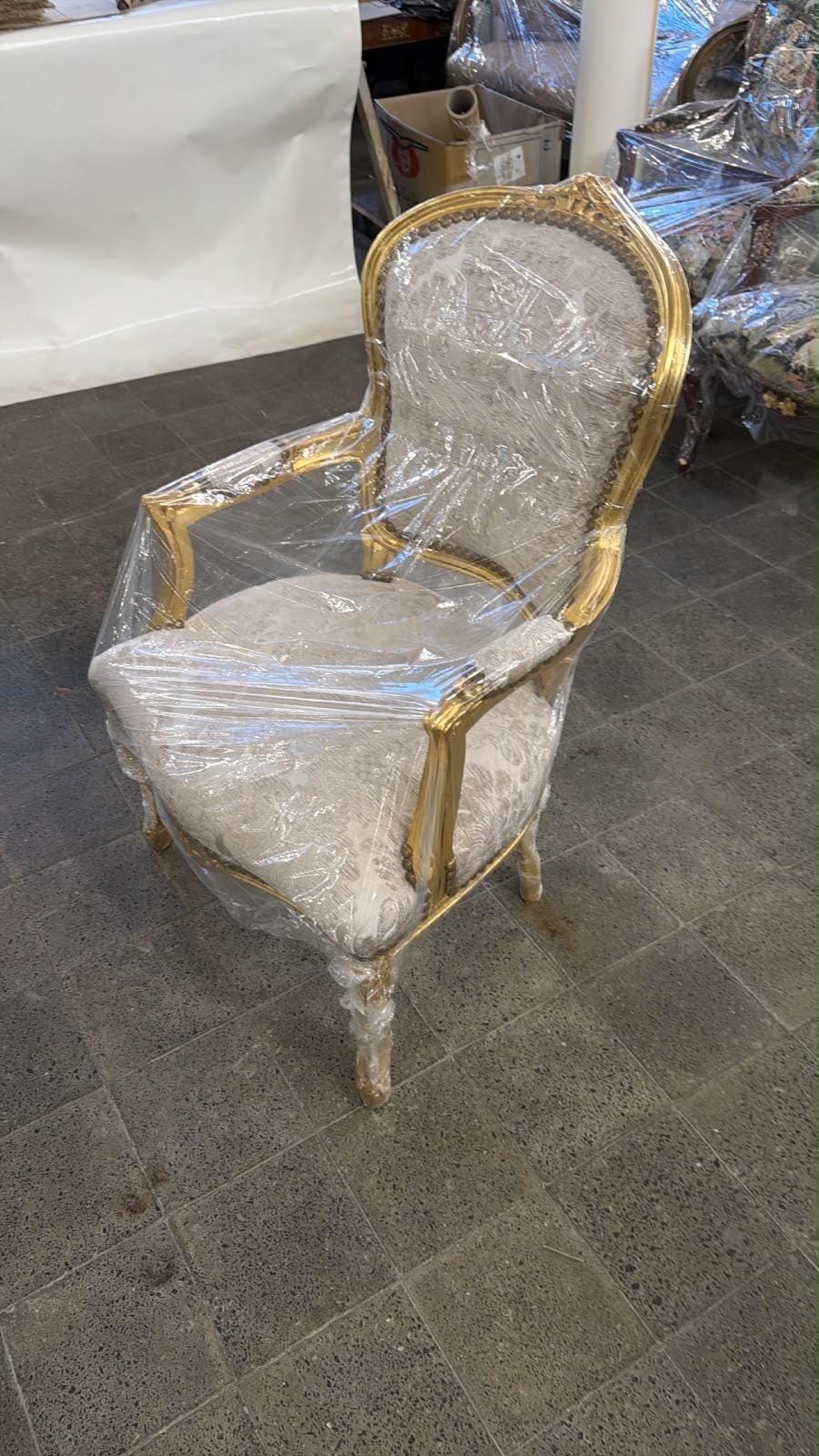 May include: An armchair with a gold-painted wooden frame and light-colored fabric upholstery featuring a subtle floral pattern. The chair has curved armrests, a rounded backrest, and is wrapped in clear plastic protective film for storage.