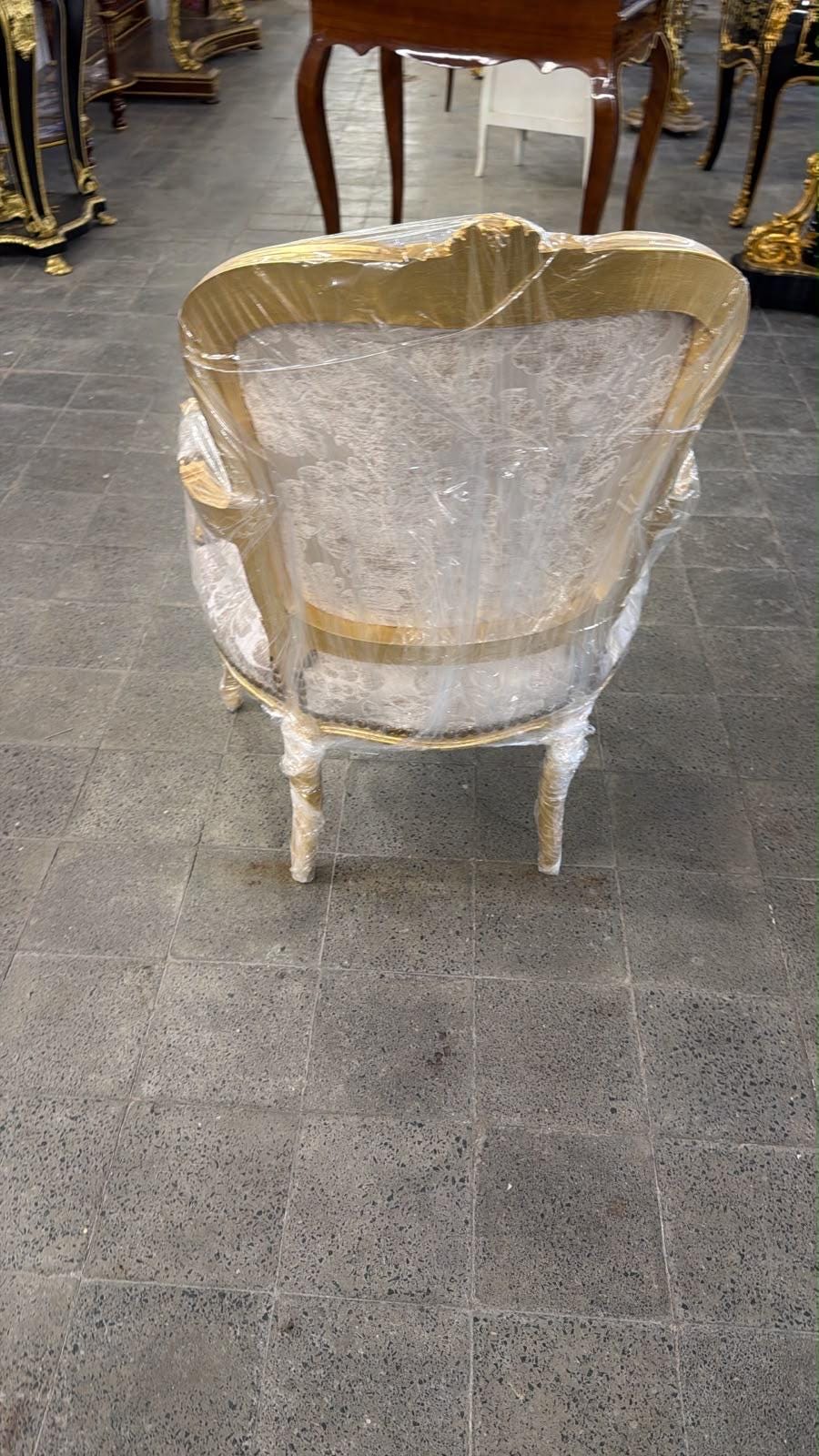 May include: An armchair with a gold-painted wooden frame and light-colored patterned upholstery, fully encased in clear protective plastic wrap. The chair features curved armrests, a rounded backrest, and four slender, slightly curved legs.