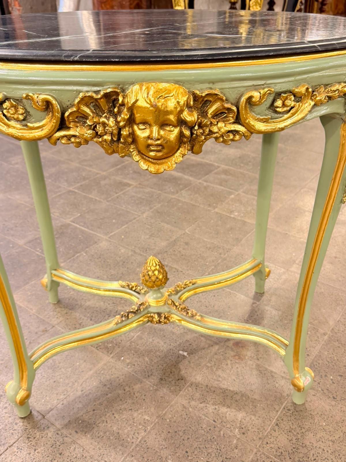 May include: A round table with a black marble top and a light green frame featuring gold-painted accents. The apron displays a central gold cherub face carving, and the four curved legs are connected by a cross-stretcher with a gold finial.