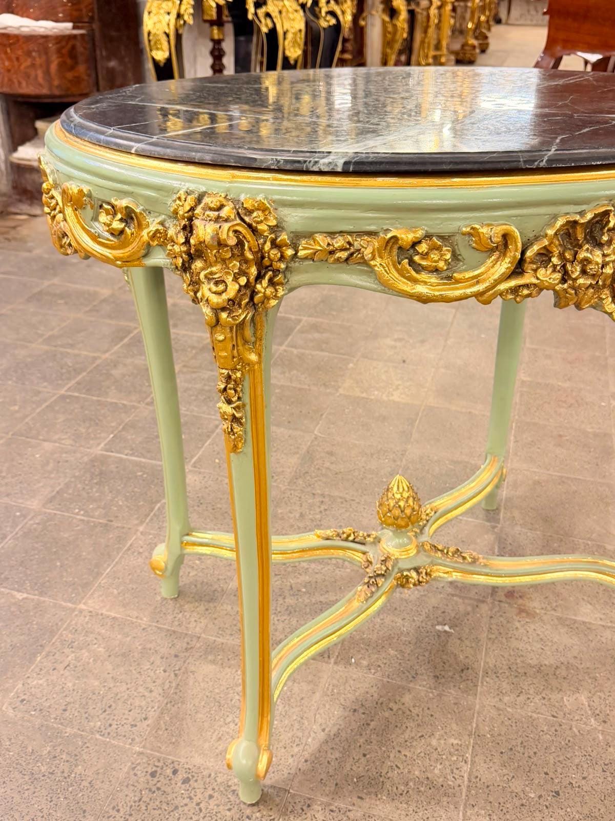 May include: A round table with a dark grey marble top featuring white veining. The table frame is painted a soft sage green with ornate gold-colored carved accents along the apron and legs. A central stretcher connects the four legs.