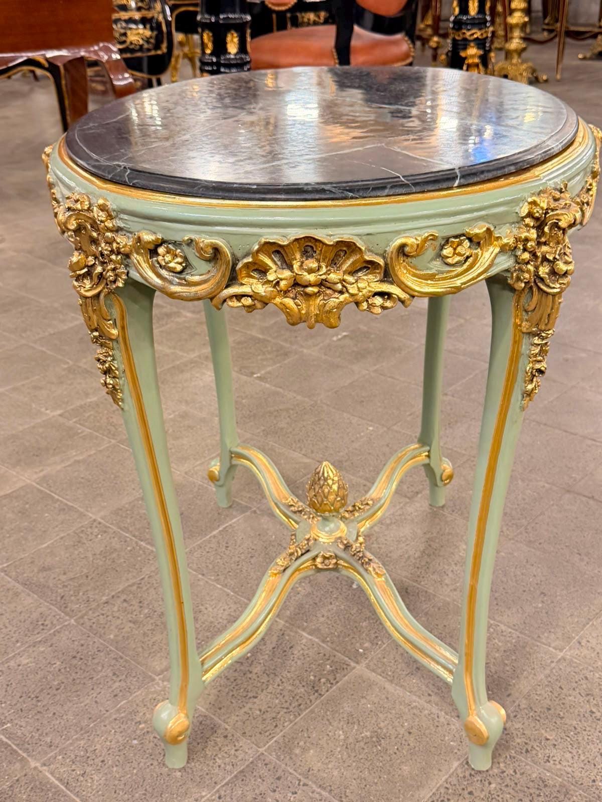 May include: A round side table with a black marble top and a sage green wooden frame. The table features ornate gold-painted carved details along the apron and legs, with a central decorative finial where the four curved legs meet at the base.