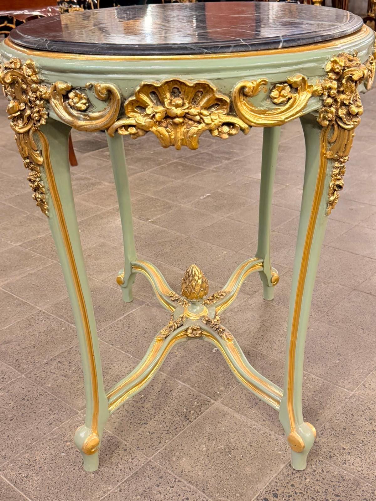 May include: A round table with a black marble top and a sage green frame featuring gold-painted ornate carvings. The table has four slender legs connected by a cross-shaped stretcher with a central decorative pinecone-shaped finial.