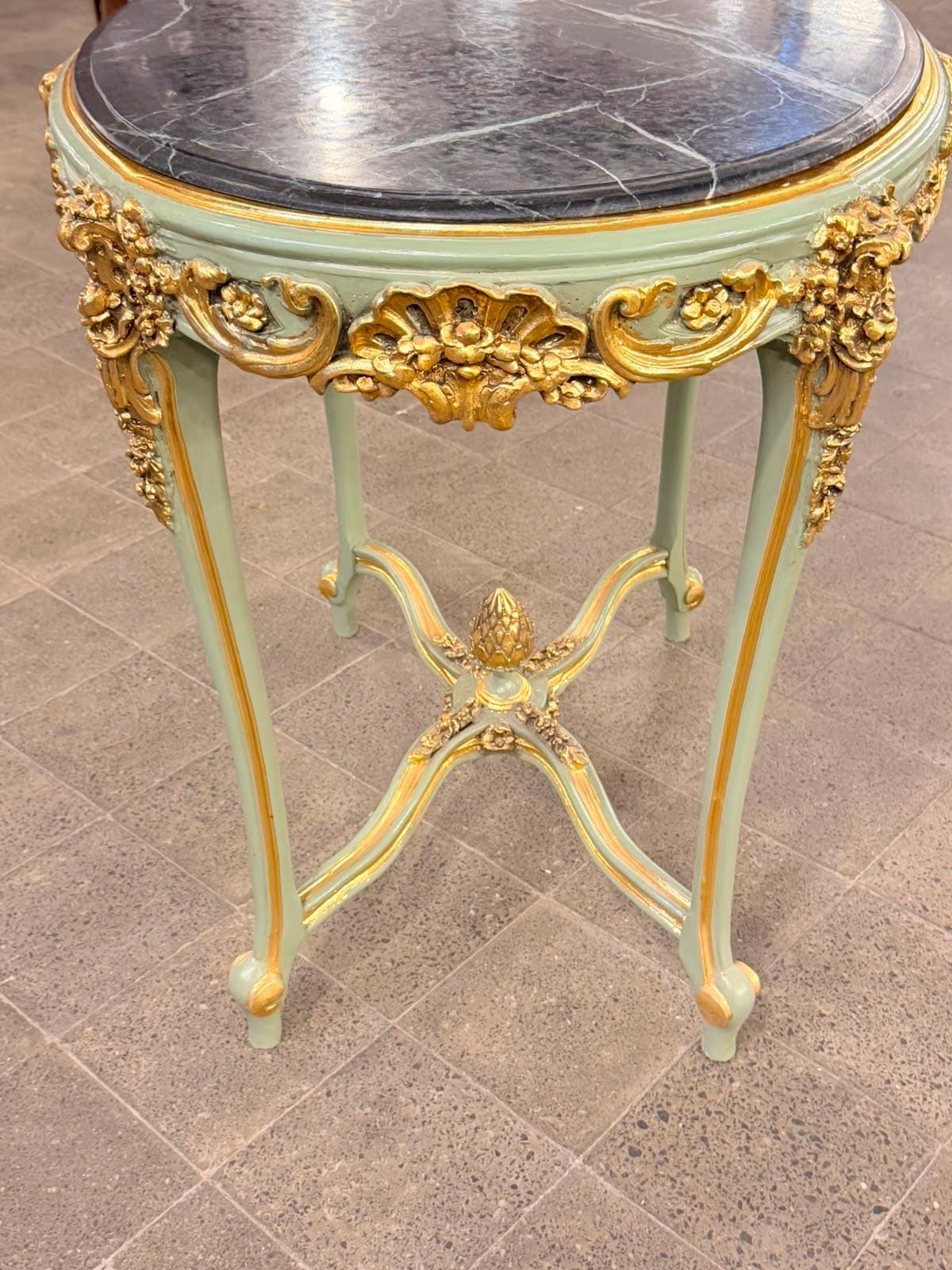 May include: A round side table featuring a black marble top with white veining. The frame is painted in a soft sage green with ornate gold-colored carved details along the apron and legs. An X-shaped stretcher connects the four curved legs.
