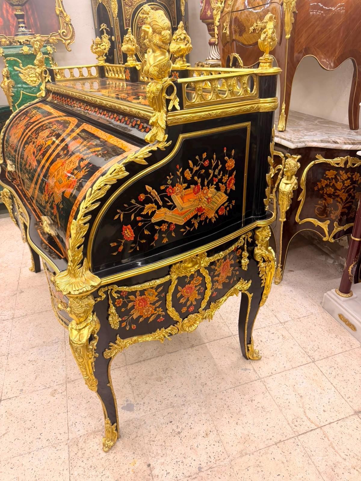 May include: A black roll-top desk featuring intricate gold-toned metal ornamentation, including ornate scrollwork and figural accents. The exterior surfaces are decorated with detailed floral and book-themed marquetry in shades of orange and red.