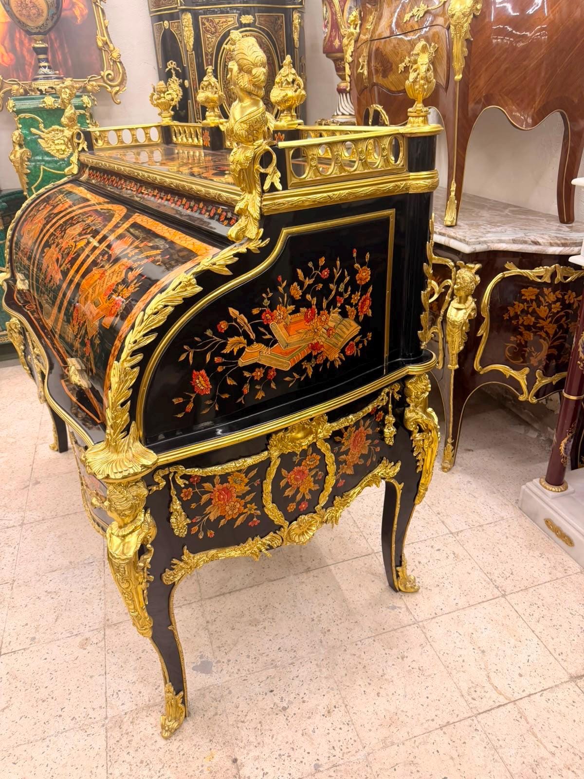 May include: A black roll-top desk featuring intricate gold-toned metal mounts and ornate floral marquetry in shades of red and orange. The piece includes a curved tambour door, cabriole legs, and decorative metal finials along the top edge.