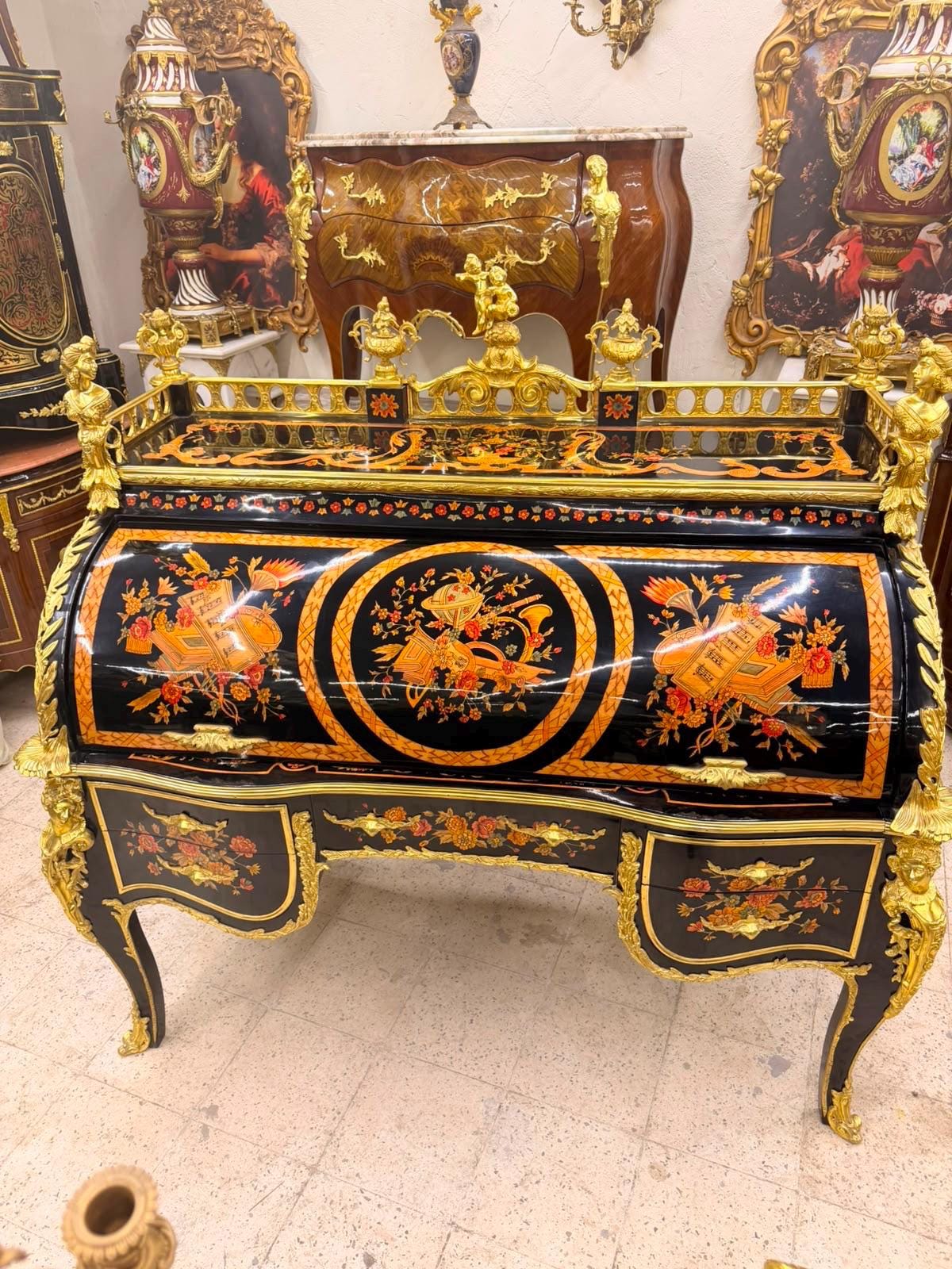 May include: A black roll-top desk featuring intricate gold ormolu mounts, floral marquetry, and a curved front. The desk includes a gallery top with gold figures, multiple drawers with floral inlays, and curved legs with ornate gold metal accents.