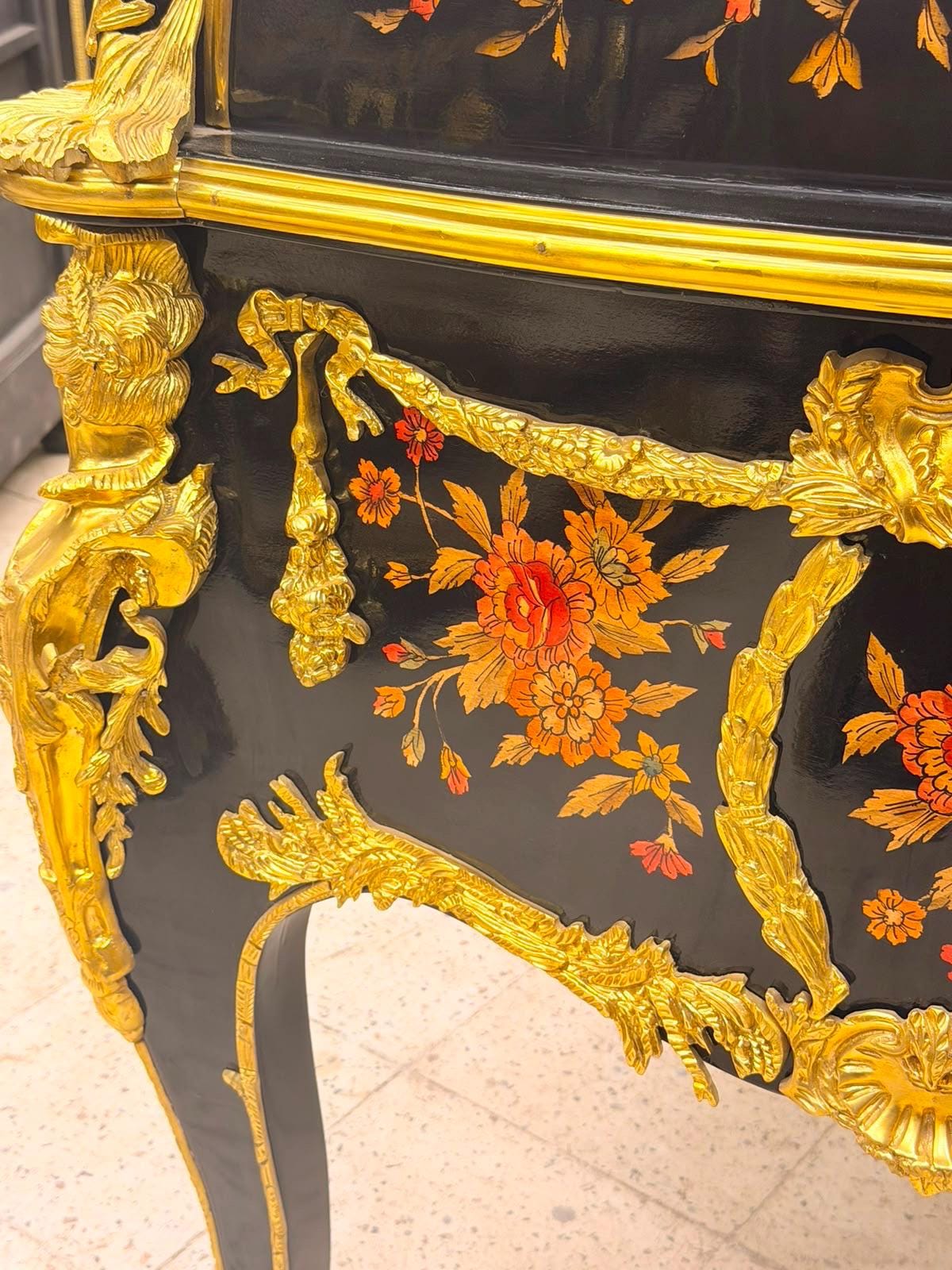 May include: A black lacquered cabinet featuring ornate gold-toned metal mounts and decorative floral motifs in shades of orange and red. The piece includes intricate relief carvings of ribbons and foliage, with a glossy finish and curved legs.