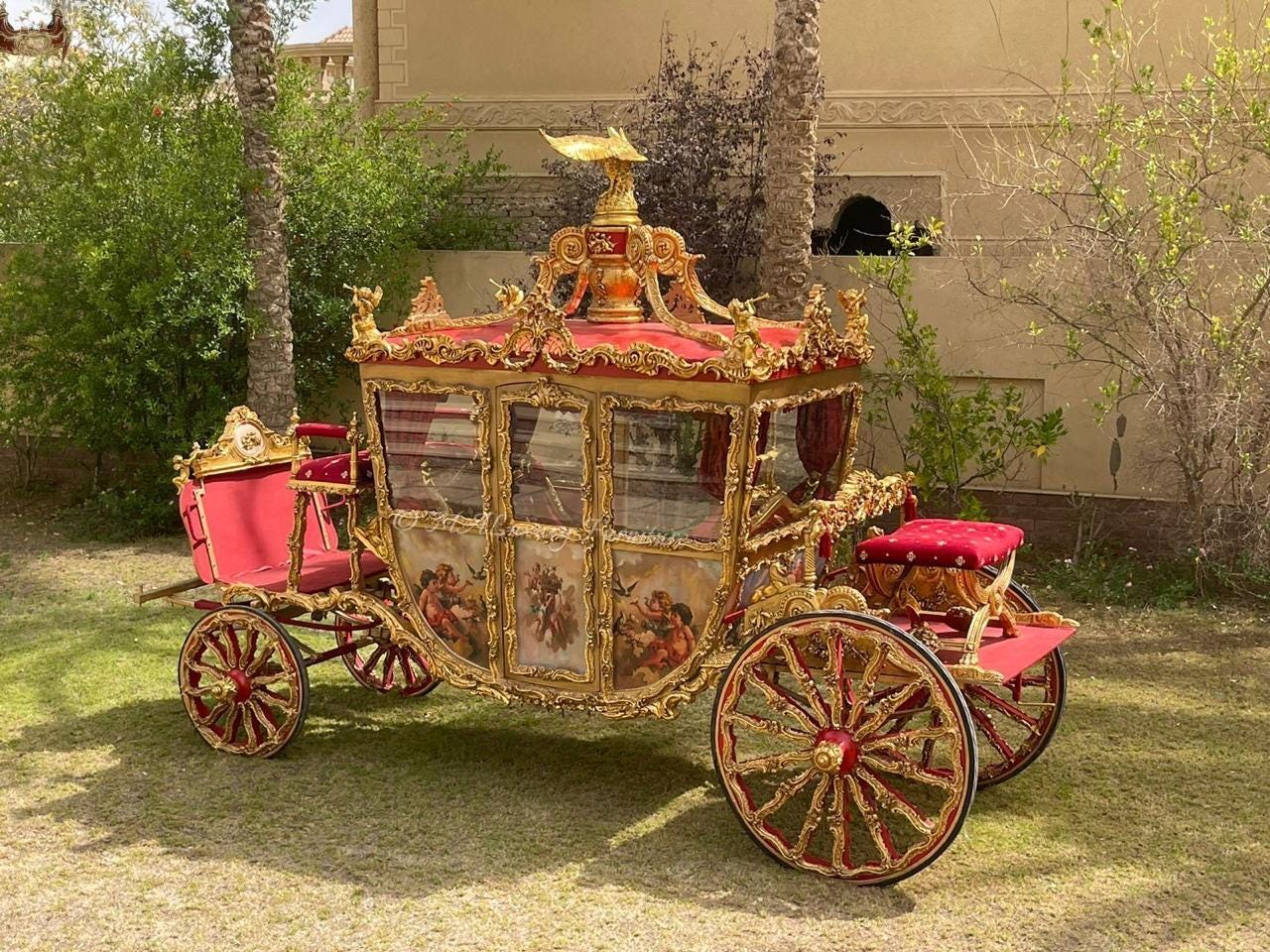 May include: A decorative horse-drawn carriage featuring a gold-painted frame with intricate scrollwork, red velvet upholstery, and painted panels depicting figures. The carriage has large spoked wheels and a golden eagle ornament on the roof.