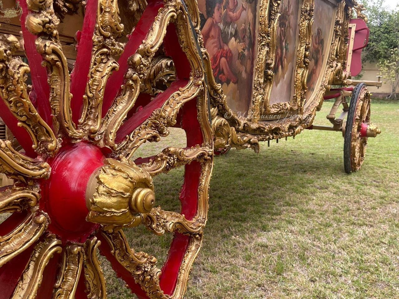 May include: A large carriage featuring a red and gold wheel with intricate carved details. The body of the carriage displays painted panels depicting figures in a classical style, with ornate gold trim throughout the structure and frame.