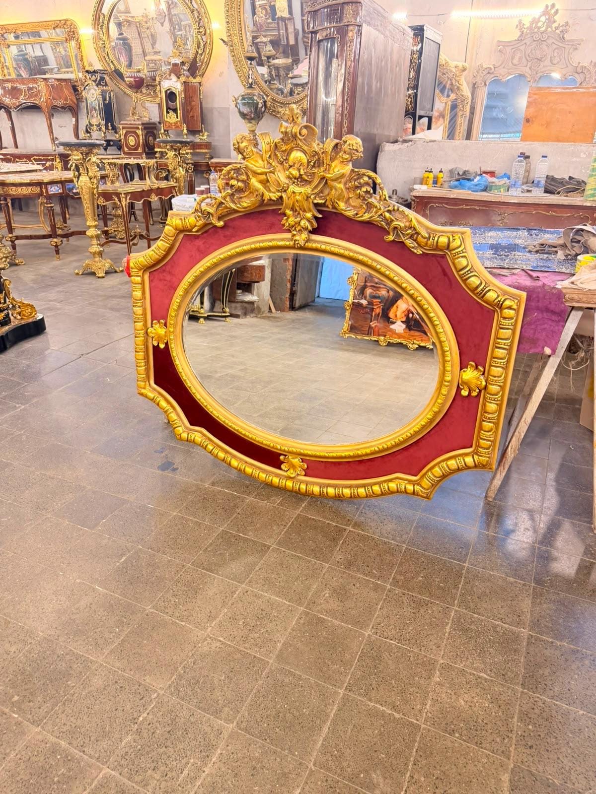 May include: An oval wall mirror featuring a wide, ornate gold-painted frame with a deep red velvet inlay. The top of the frame is decorated with sculpted cherub figures and intricate floral carvings, while the edges display repeating geometric patterns.