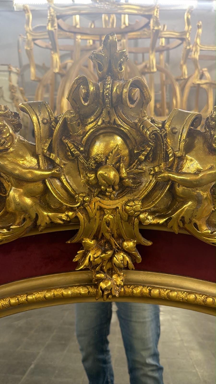 May include: A gold-finished mirror frame featuring ornate carvings of two cherubs flanking a central crest with floral and scrollwork details. The frame includes a deep red velvet border and a beaded inner edge, showcasing intricate relief work.