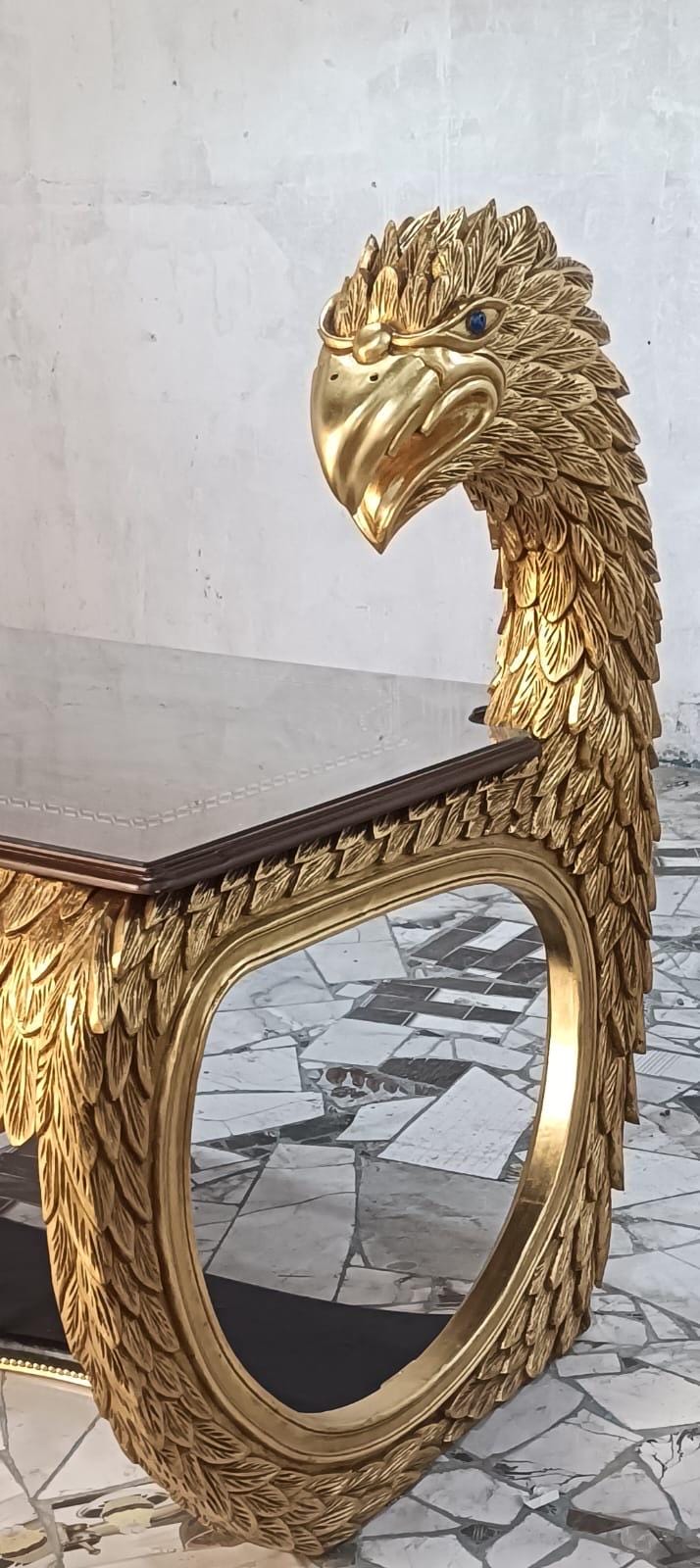 May include: A gold-colored table featuring a carved eagle head and neck as a support structure. The eagle has detailed feather carvings and a blue eye. The table has a rectangular top with a dark surface and a smooth, oval-shaped base frame.