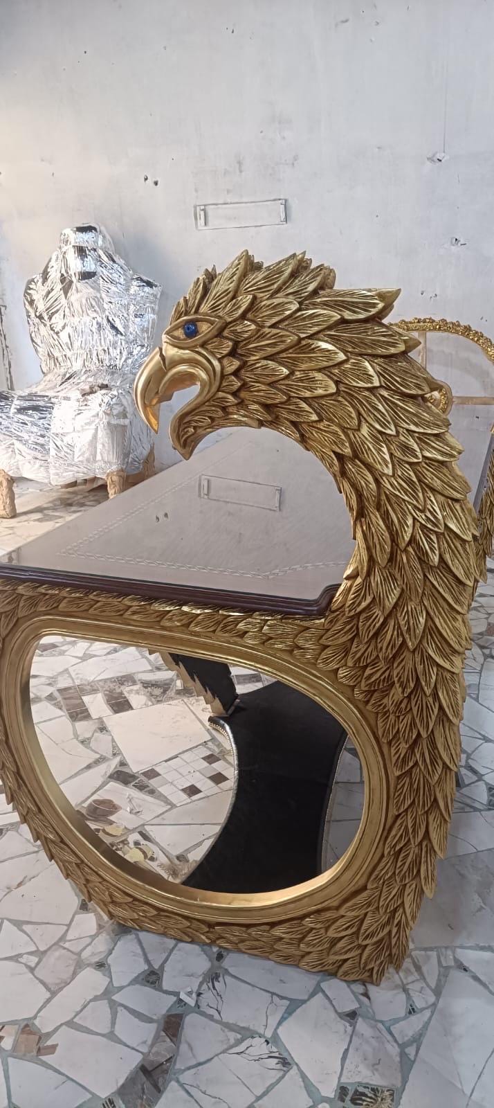 May include: A gold-colored desk featuring a large, carved eagle head and neck as the primary support structure. The eagle has detailed feather textures and a single blue eye. The desk surface is rectangular with a dark, smooth top and a gold frame.