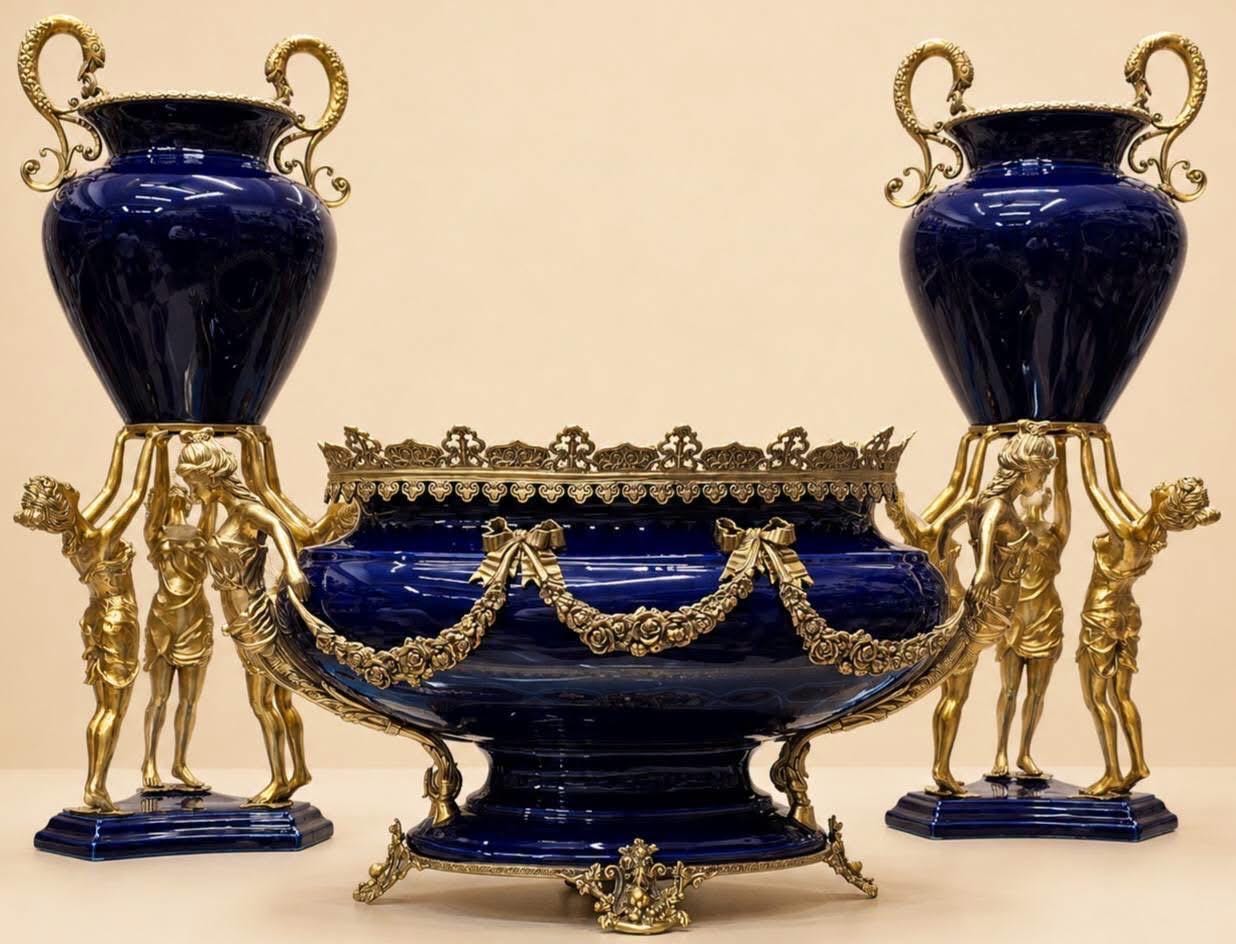 May include: A set of two deep blue porcelain urns with gold-toned metal handles and bases, each supported by three gold-toned figures, alongside a matching deep blue porcelain centerpiece bowl featuring gold-toned metal floral garlands and trim.