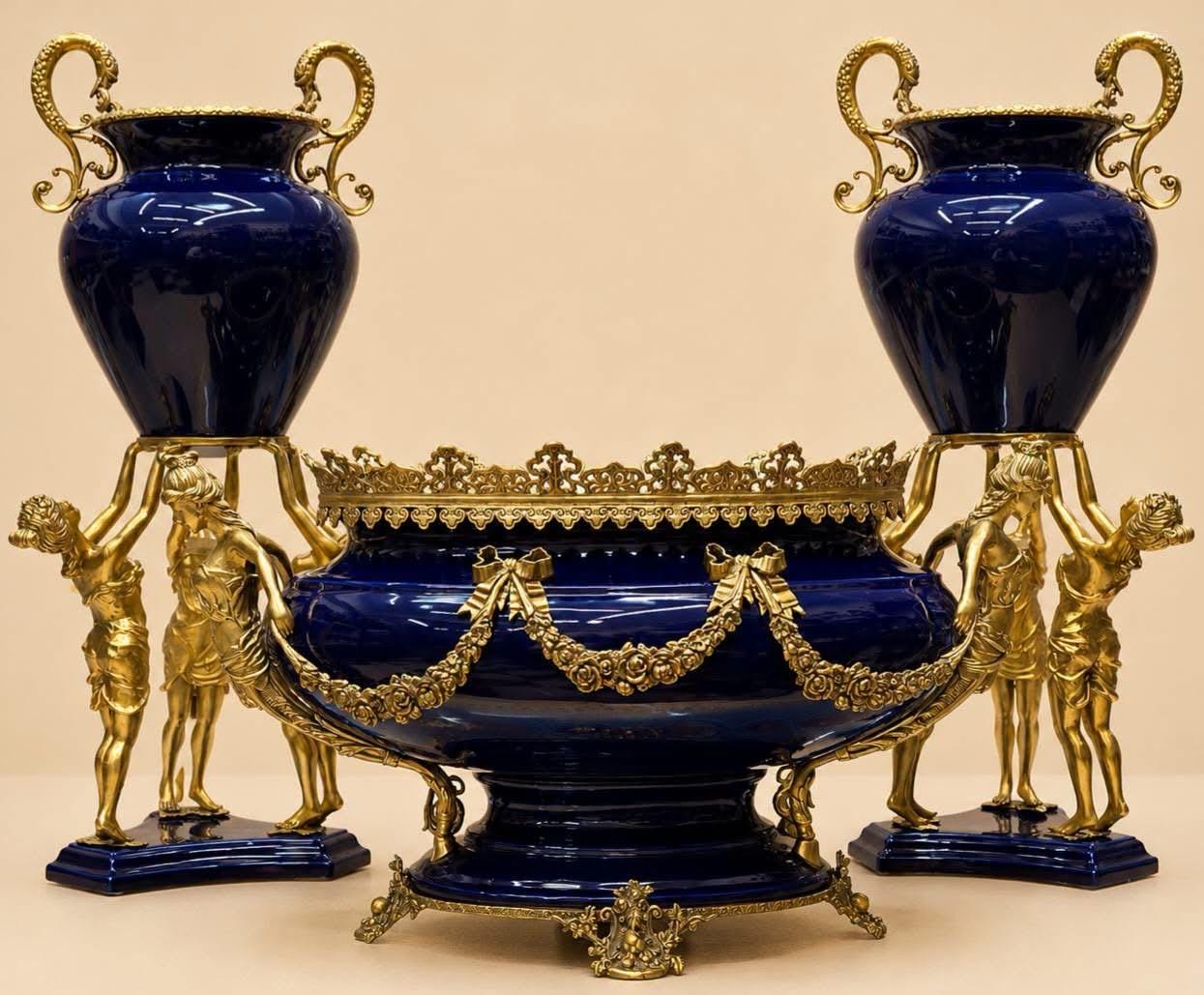 May include: A set consisting of a large central bowl and two matching urns, all crafted from deep blue ceramic with ornate gold-toned metal mounts. The bowl features a decorative rim and floral swags, while the urns are supported by gold figures.