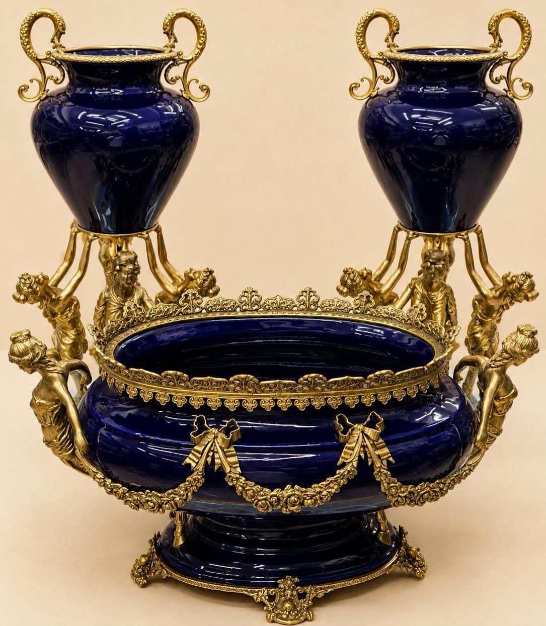 May include: A deep blue porcelain centerpiece bowl and two matching urns featuring ornate gold-toned metal mounts. The bowl has a decorative rim and bow-and-swag accents, while the urns are supported by sculpted figures on gold bases.
